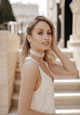 Stylish blonde grey-eyed woman with beautiful makeup in pearl necklace and white dress looks into camera and smiles outside.