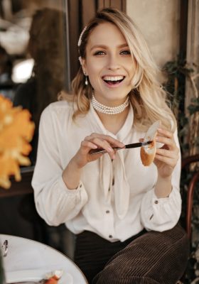 Happy cheerful blonde woman in stylish white blouse and dark velvet pants laughs, looks into camera and spreads paste on piece of bread in street cafe.